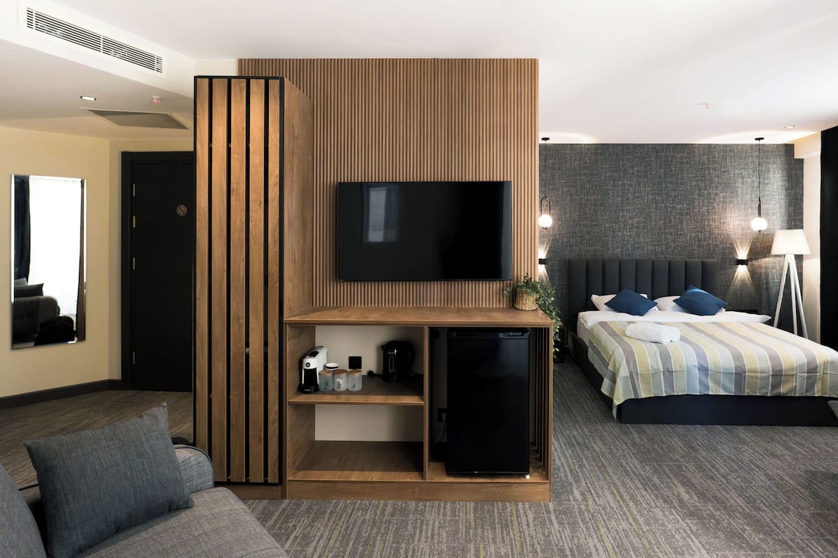 Une chambre d’hôtel moderne dotée d’un grand téléviseur à écran plat fixé sur un mur en panneaux de bois, d’un lit avec une literie à rayures, d’une table de chevet avec une lampe et d’un petit coin salon avec un canapé et des coussins.
