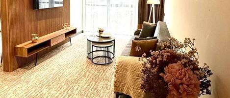 A modern hotel room with a flat-screen TV mounted on a wooden panel wall, a sofa, a small round coffee table, and a dining table with a vase of flowers.