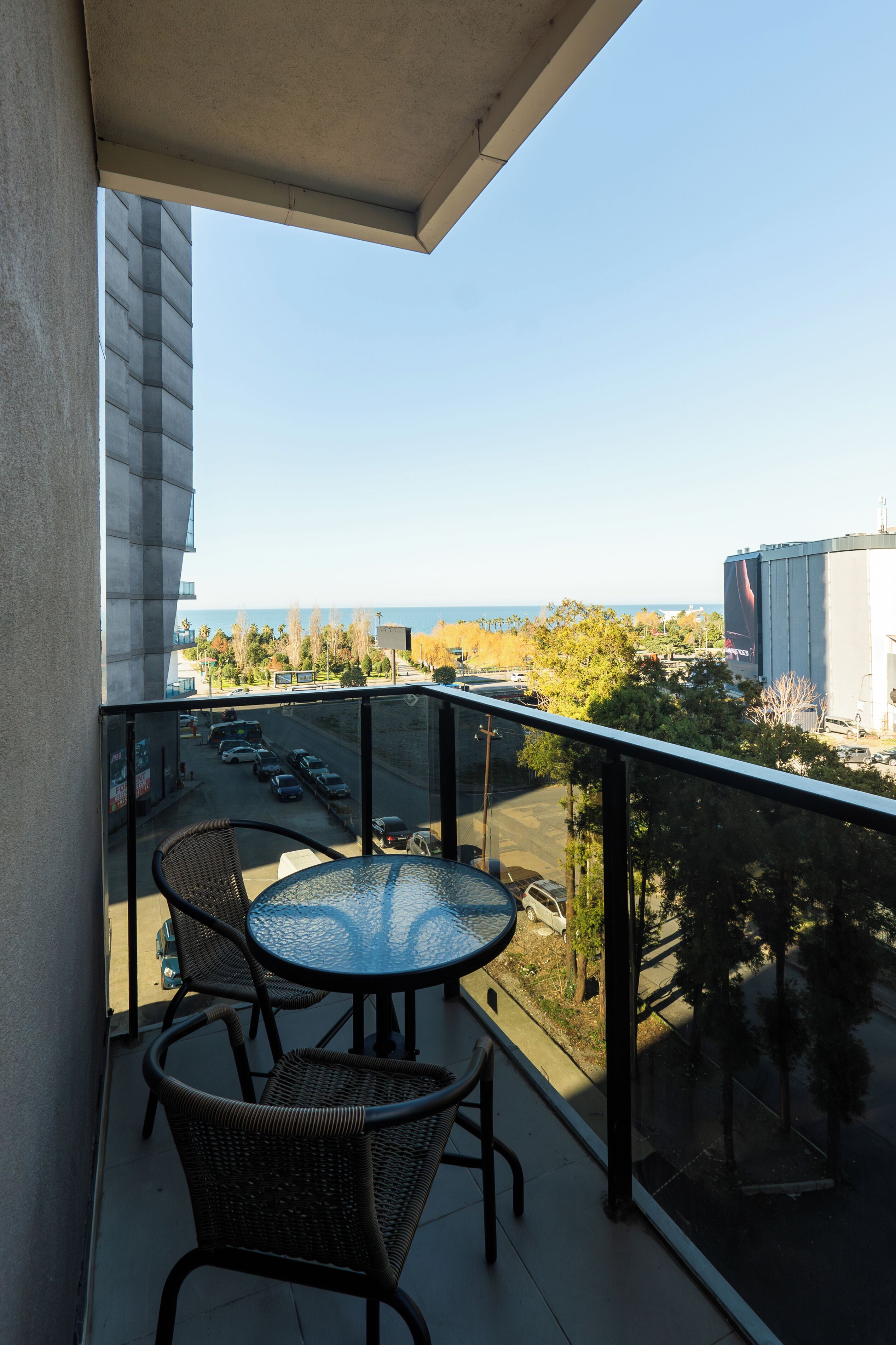 A balcony with a glass table and wicker chairs, overlooking a parking lot and a distant view of the sea.