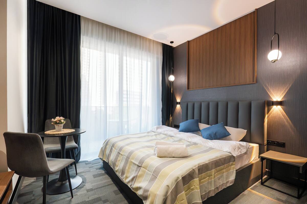 Une chambre d’hôtel avec un lit, une table de chevet, une chaise et une petite table sur laquelle est posé un vase de fleurs.