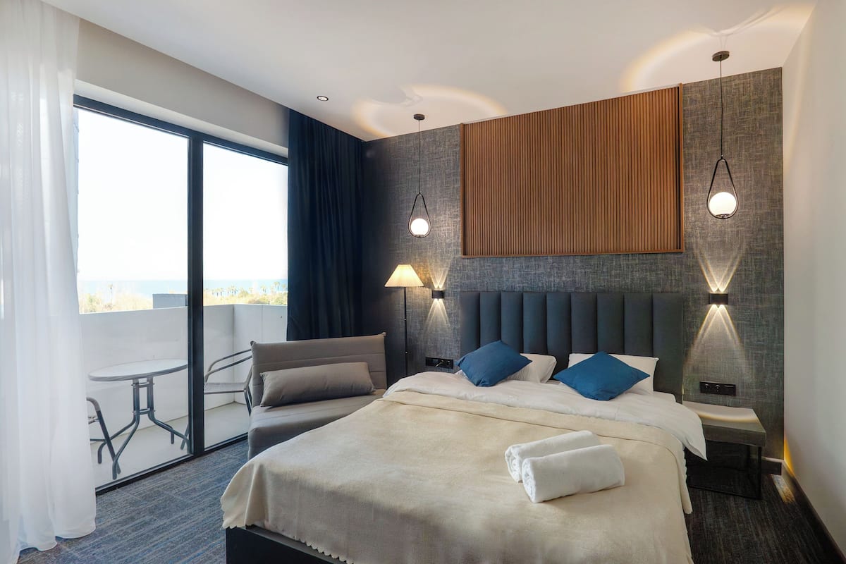 Une chambre d’hôtel moderne dotée d’un grand lit, de deux tables de chevet, d’une commode, d’un balcon avec une table et des chaises, et offrant une vue sur la ville.