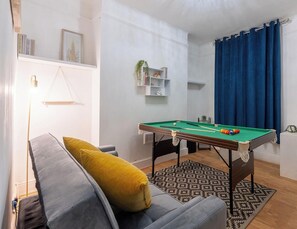 Une pièce avec une table de billard, un canapé agrémenté d’un coussin jaune, une bibliothèque et une fenêtre avec des rideaux bleus.