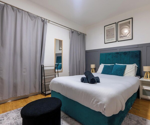 Une chambre à coucher bien rangée, avec un lit, une table de chevet, une lampe, un miroir et une fenêtre avec des rideaux.