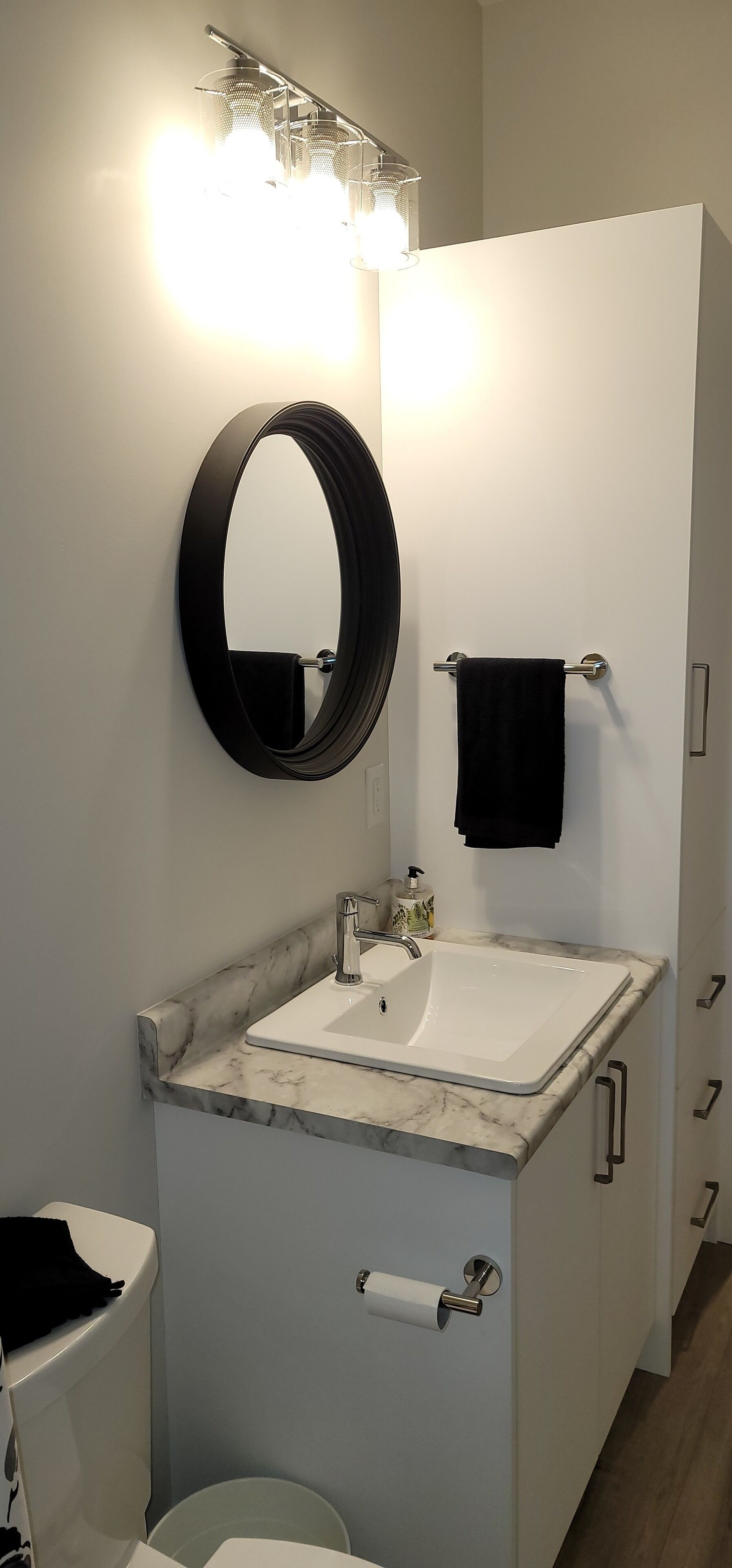 Une salle de bain avec un lavabo blanc, un miroir rond, une serviette noire et des toilettes.