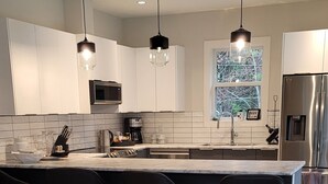 A modern kitchen with a large island, white cabinetry, and stainless steel appliances. There is a beige leather sofa with a plaid pillow in the foreground. - Double D Ranch Guest House (Courtenay)