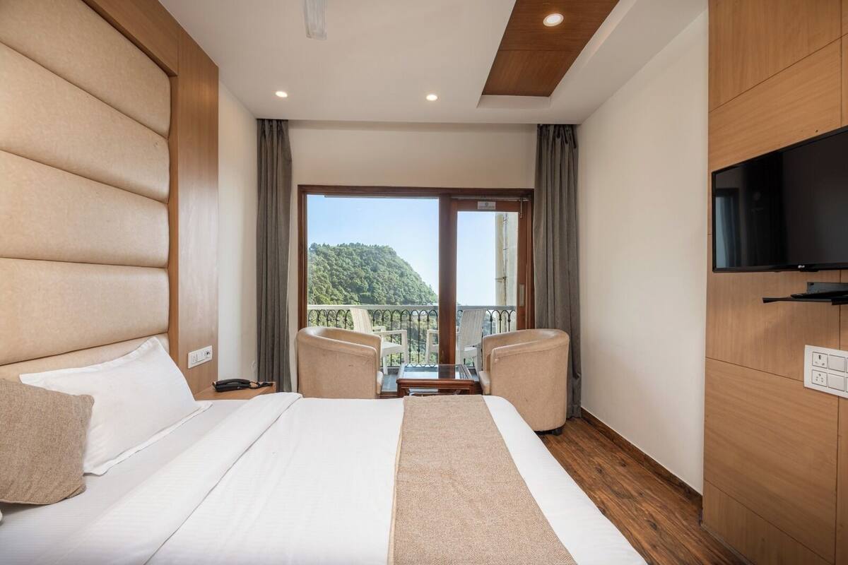 A hotel room with a large bed, two armchairs, a TV, and a balcony with a view of a forested hill.