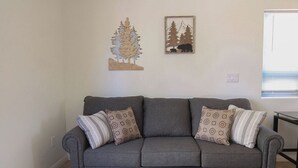 Un sofá gris con cojines estampados, un adorno de árbol de madera y un cuadro en la pared con un oso y árboles.