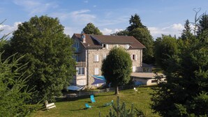 A two-story stone building with a sloped roof, surrounded by trees and a grassy lawn with outdoor furniture. - Group gîte with single rooms, no dormitories, in the heart of nature. (Le Chambon-sur-Lignon)