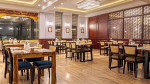A spacious dining area with wooden tables and chairs, large windows with decorative screens, and a chandelier. - Hotel Samaya (Leh)