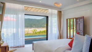 A hotel room with a large bed, a desk, and a view of a beach and mountains through the window. - Chaloklum Residences by Kupu Kupu Phangan (Ko Pha-ngan)