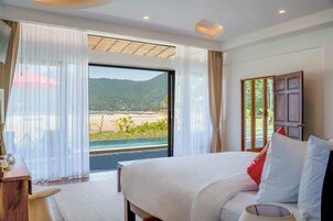 Habitación de hotel con una cama grande, un escritorio y vistas a la playa y montañas desde la ventana.