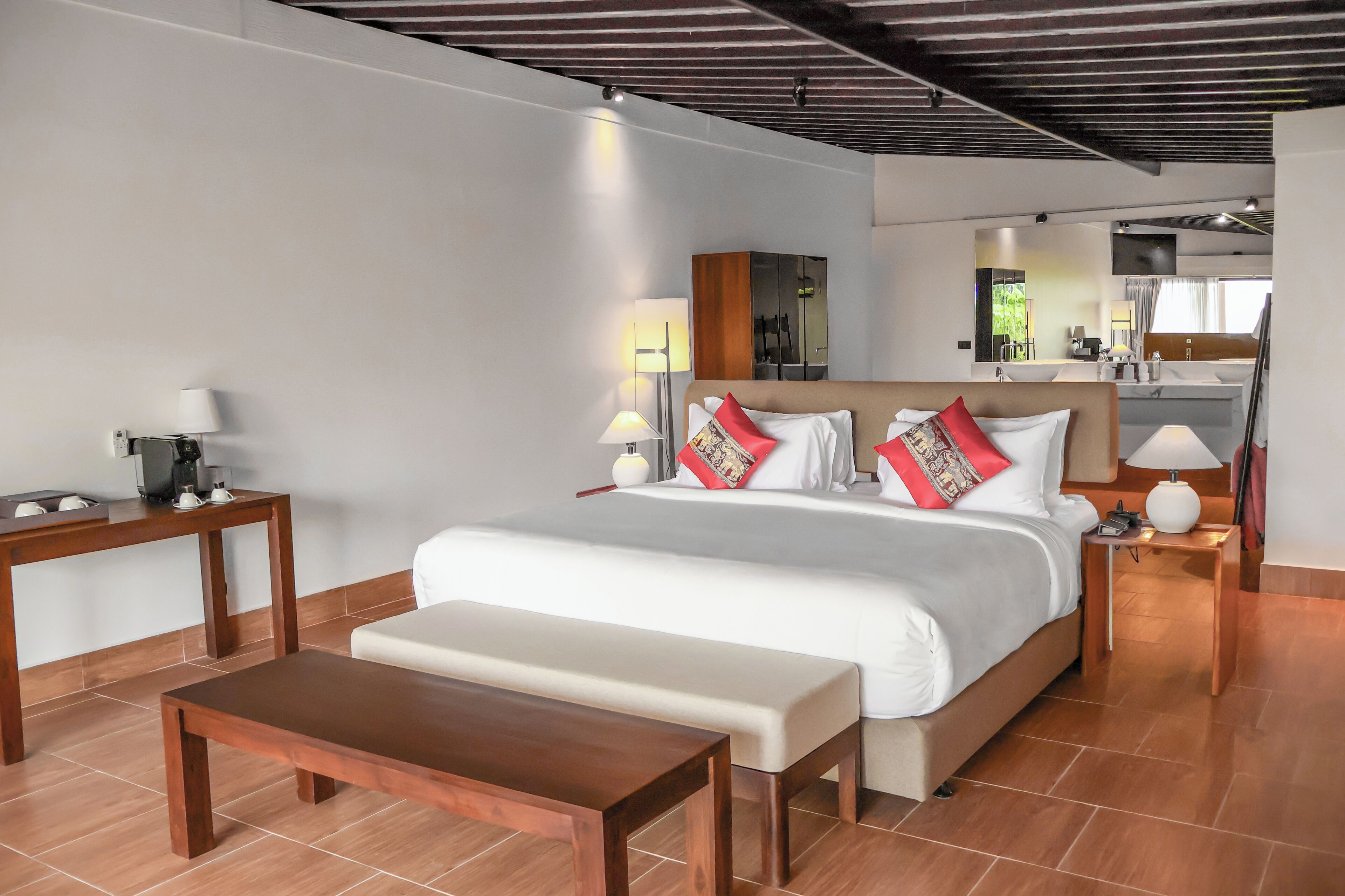 A modern hotel room with a large bed, a wooden bench, a small table, and a kitchenette in the background.