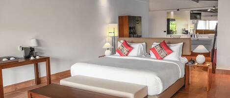 A modern hotel room with a large bed, a wooden bench, a small table, and a kitchenette in the background.