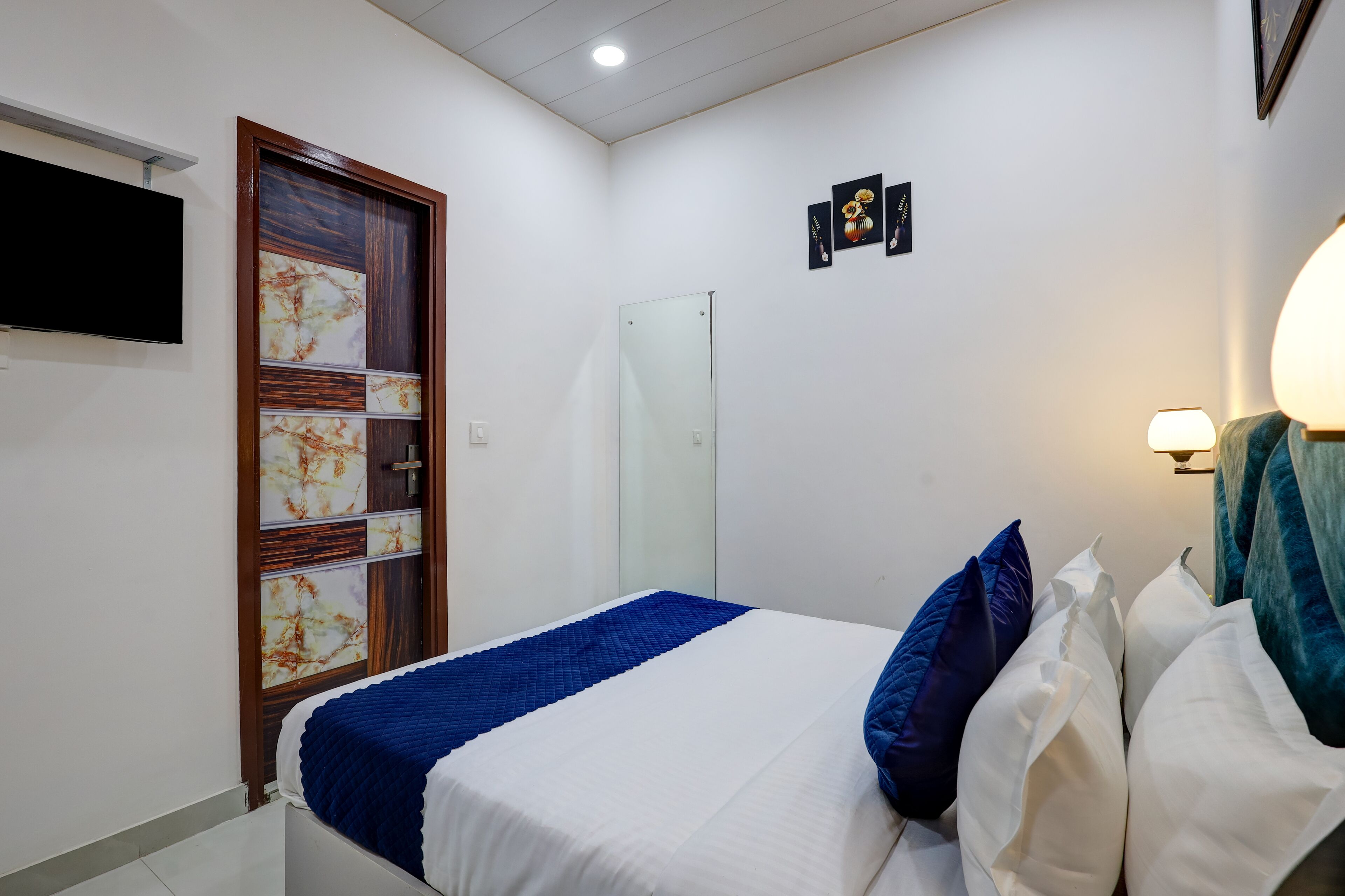 A bedroom with a bed, a TV mounted on the wall, a door with a decorative panel, and a wall-mounted lamp.