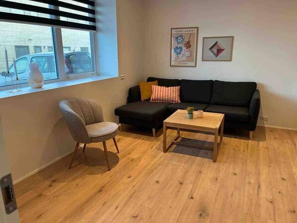 A living room with a black sofa, a wooden coffee table, and a grey armchair. There is a window with blinds, a poster on the wall, and a vase on the windowsill. - Cozy Ocean View Apartment in Hoyvík, Faroe Islands (Hoyvík)