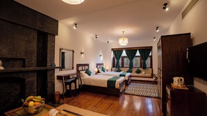 A hotel room with a wooden floor, two beds with green bedding, a television, a mirror, a small table with a fruit bowl, and a fireplace.
