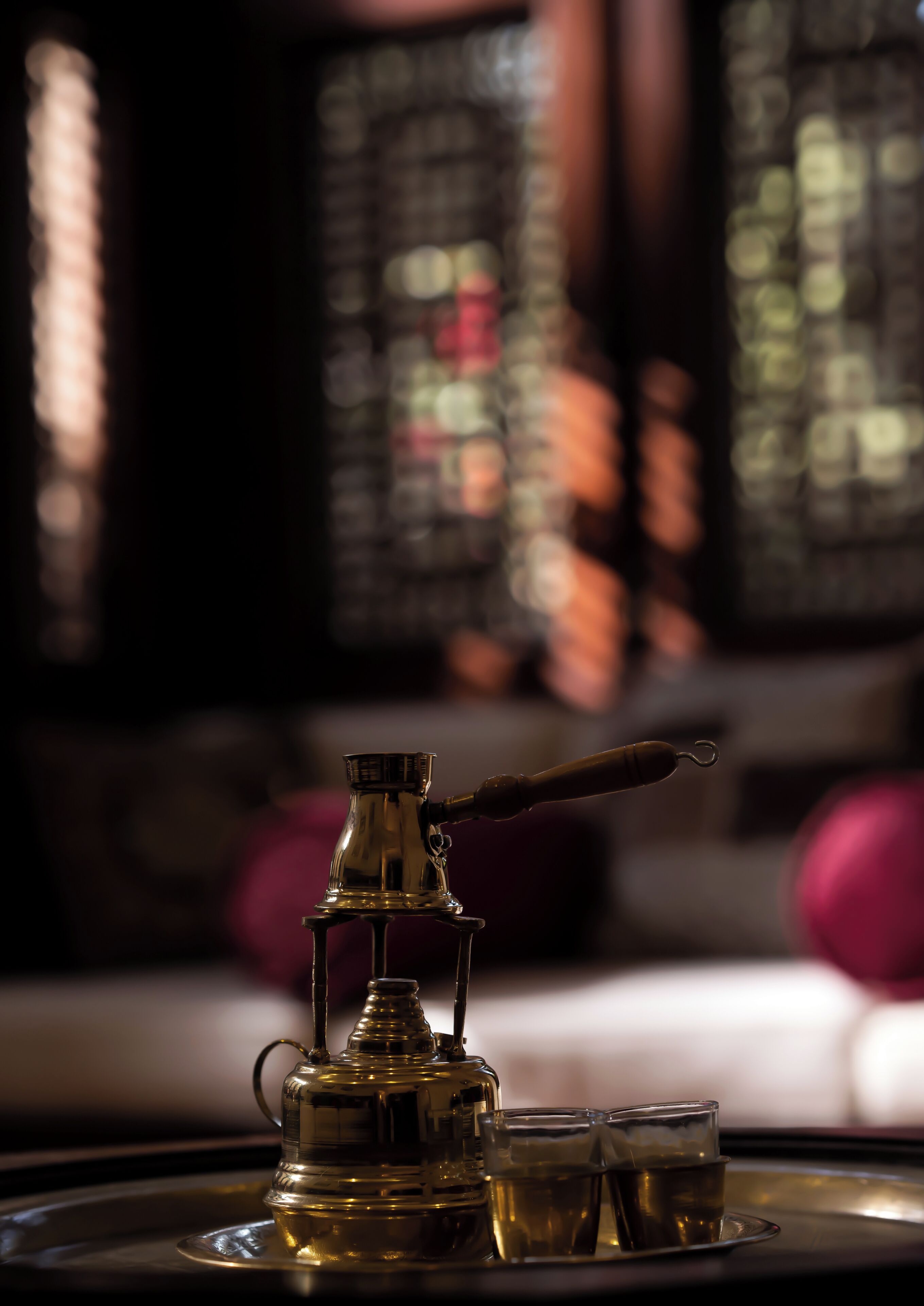 A brass teapot and three glasses on a tray, with a blurred background of a room with a window and a couch.
