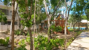 Un terrain de jeux avec des balançoires et un toboggan, entouré d’arbres et avec une piscine en arrière-plan.