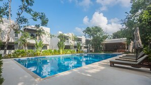 Une piscine entourée de verdure, avec un espace de détente et des bâtiments modernes en arrière-plan.