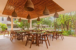 Un área de comedor al aire libre con mesas y sillas redondas, rodeada de abundante vegetación y una piscina.
