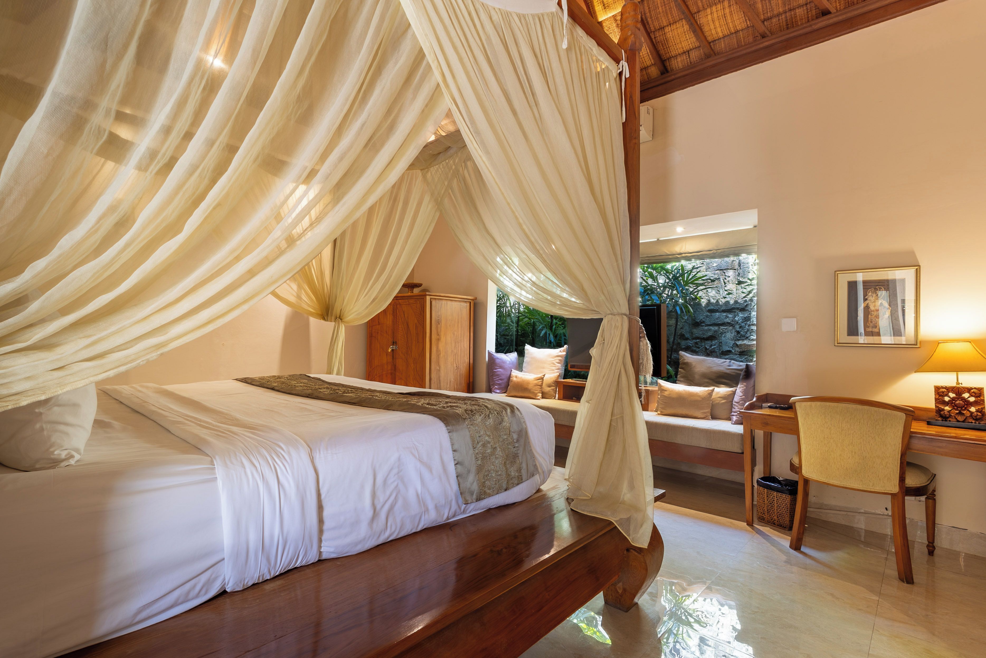 A bedroom with a four-poster bed, a wooden desk, a chair, and a view of greenery outside.