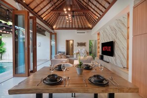 A dining area with a wooden table set for two, a TV mounted on a marble wall, and a pool visible through glass doors. - Jepun Villa by Betterplace (Seminyak)