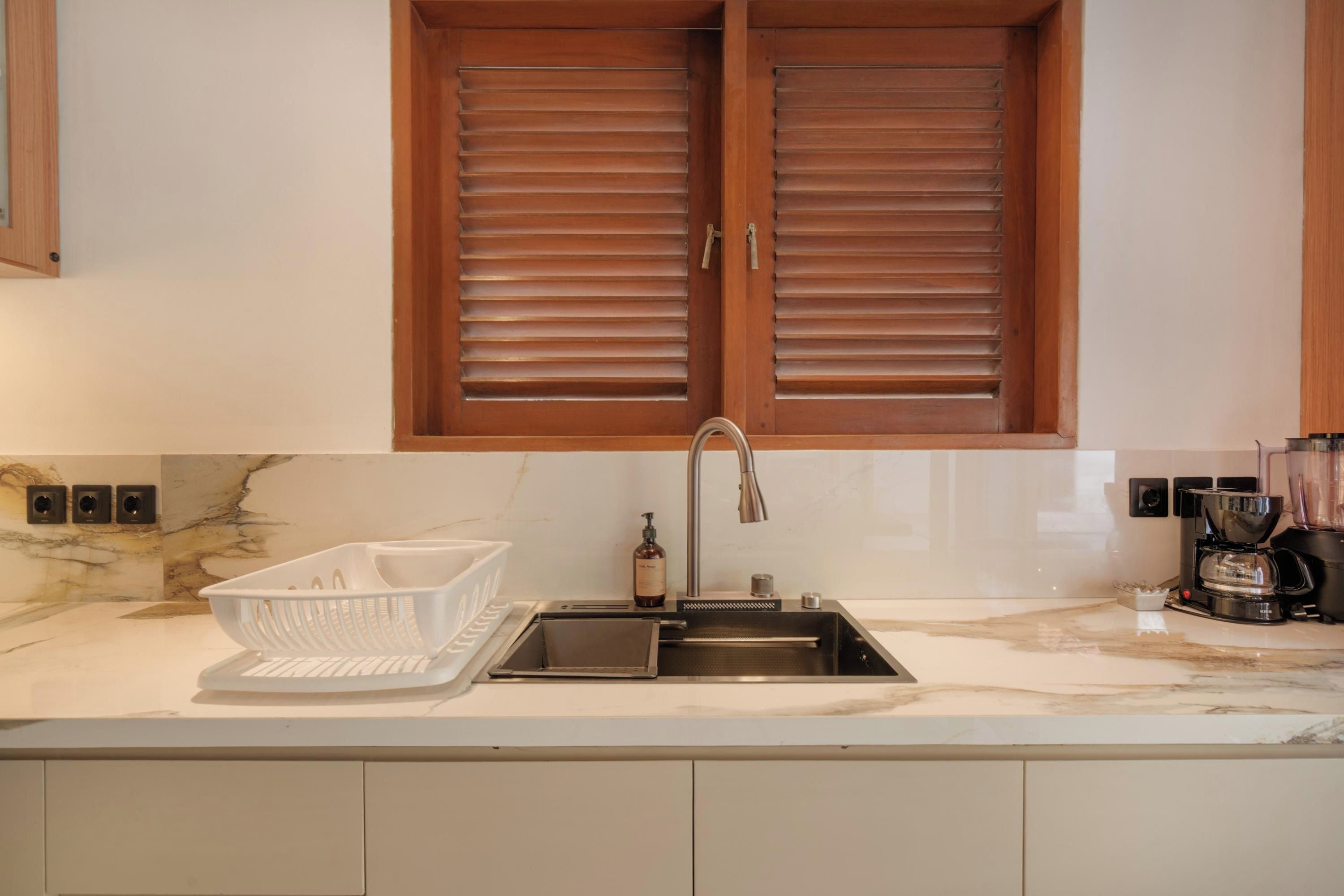 A kitchen with a marble countertop, a stainless steel sink, a white basket, and a bottle of hand soap.