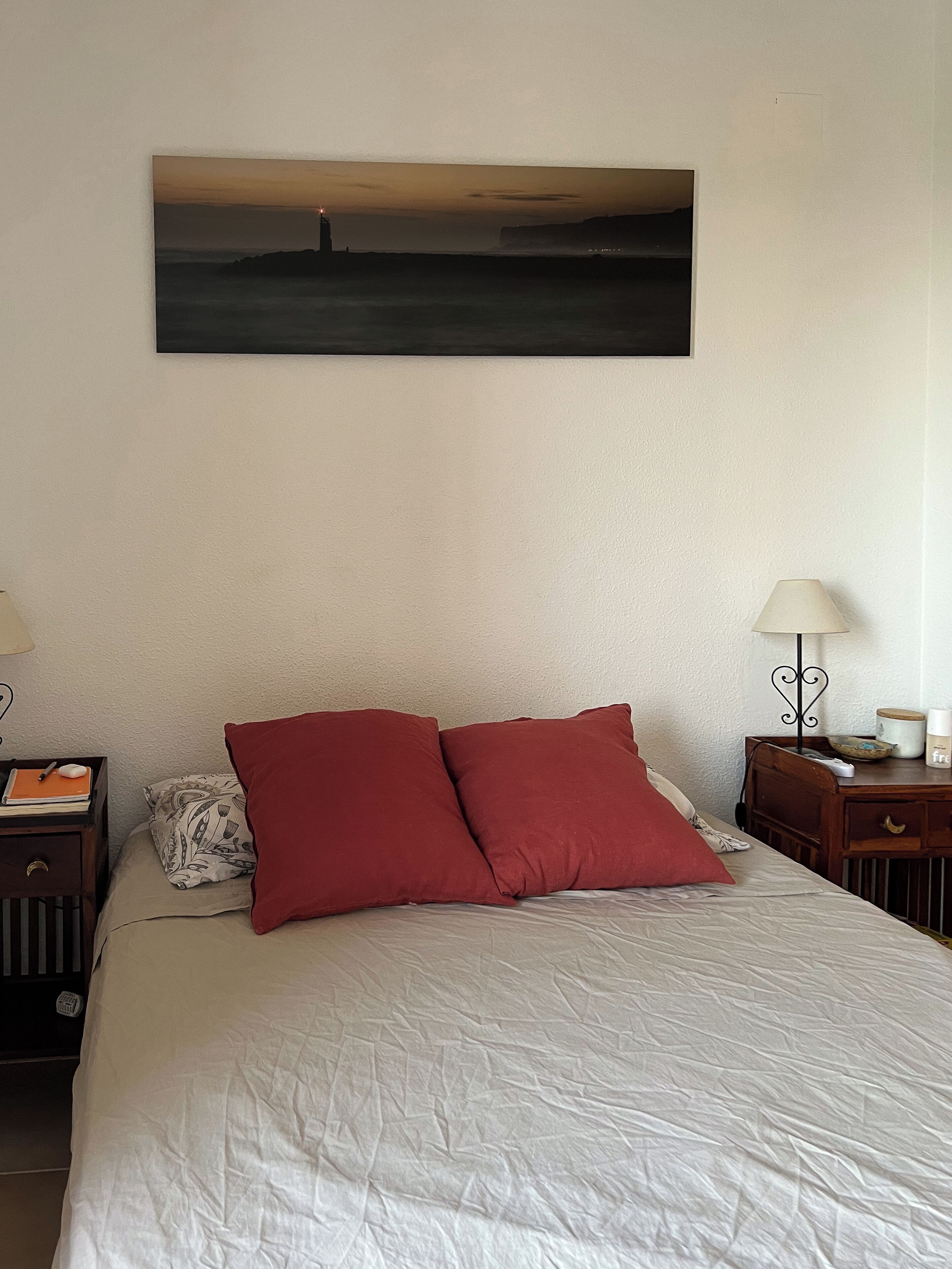 A neatly made bed with two red pillows, a white comforter, and a painting of a landscape above it. There are two bedside tables with lamps and a small nightstand with a lamp.