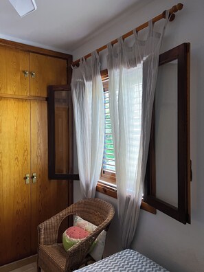 A room with a wooden wardrobe, a wicker chair with a watermelon pillow, a small bed with a patterned bedspread, and sheer curtains over a window with blinds.