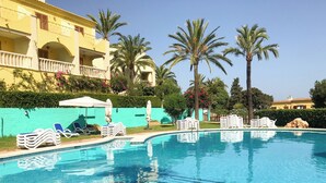 A clear swimming pool with lounge chairs and umbrellas, surrounded by palm trees and a building with balconies.