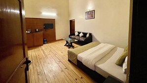 A bedroom with a wooden floor, a bed with pillows, a desk with a chair, a television on the wall, and a framed picture. - Shri Shail Inn (Dhari)