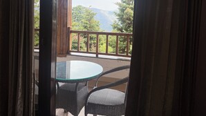A room with a glass table and wicker chairs, a wooden door, and a view of a balcony with a railing and greenery. - Shri Shail Inn (Dhari)