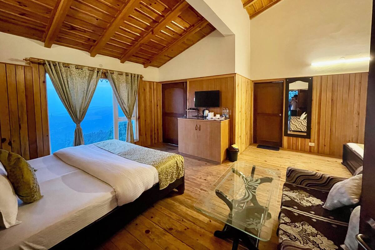 A room with a bed, a sofa, a glass table, a TV, and a view of the outdoors.