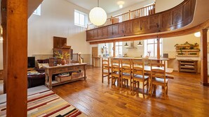 Amplia sala de estar con una mesa y sillas de madera, un techo alto con una lámpara grande, y una cocina al fondo.
