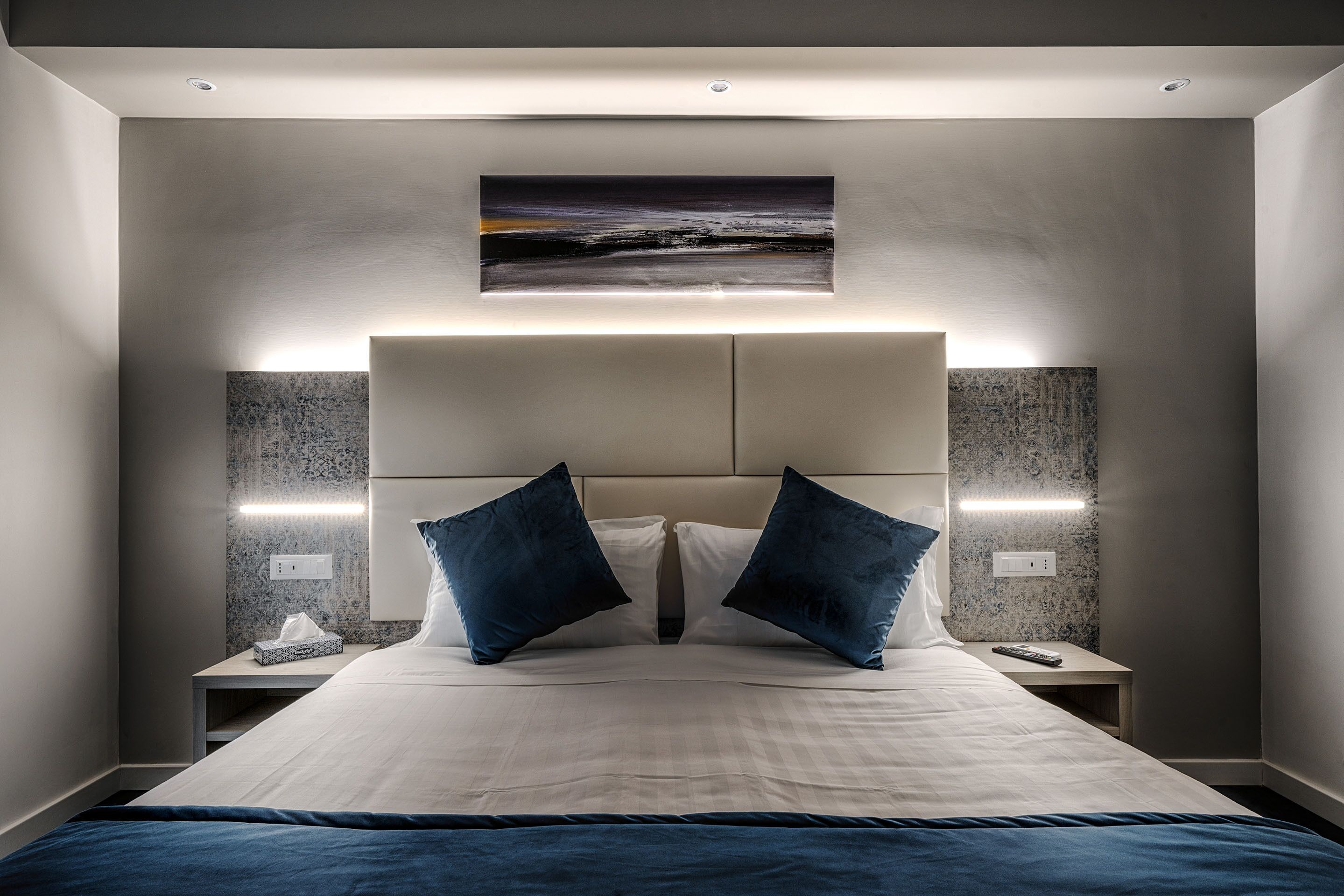 A modern hotel room with a large bed, two nightstands, a headboard with integrated lighting, and a painting above the bed.