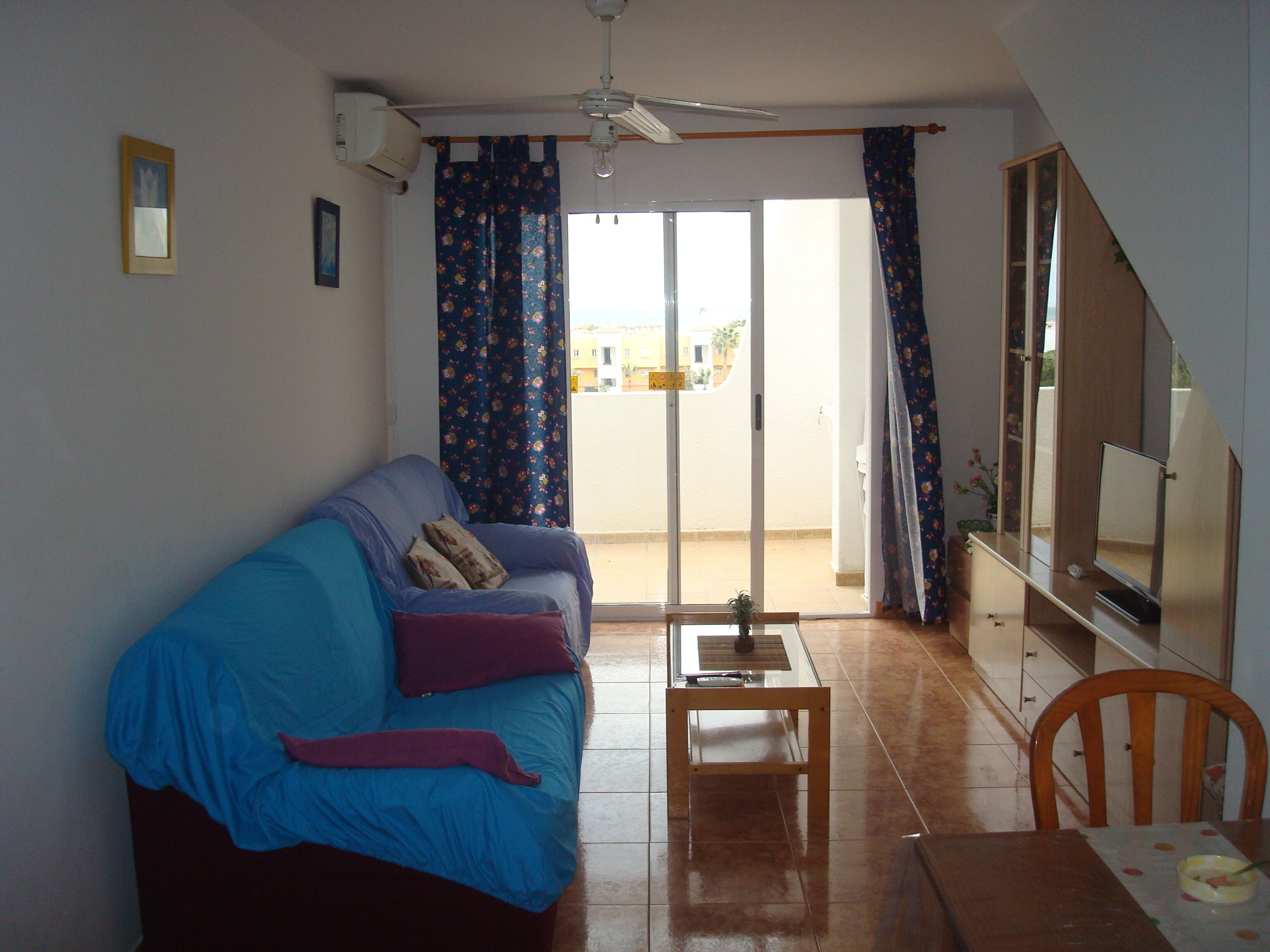Uma sala de estar com sofá, mesa de centro, televisão e varanda com cortinas.