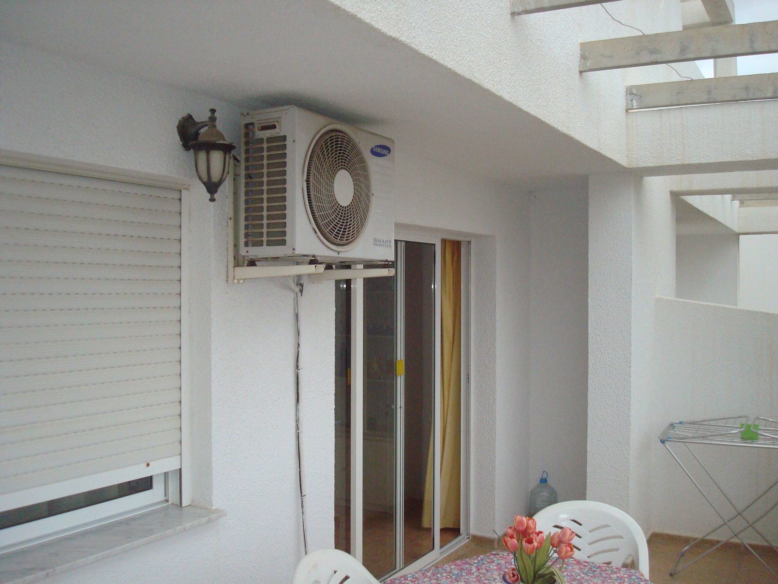 Een kamer met een witte muur, een gesloten raam met zonwering, een tafel met stoelen, een bijzettafeltje met een bloevas en een airconditioner aan de muur.