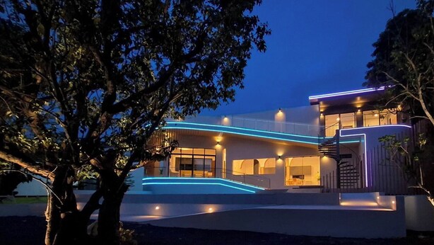 A modern house with a curved facade, illuminated by blue and purple lights, surrounded by trees. - Celine House by Craft Village (Koh Samui)