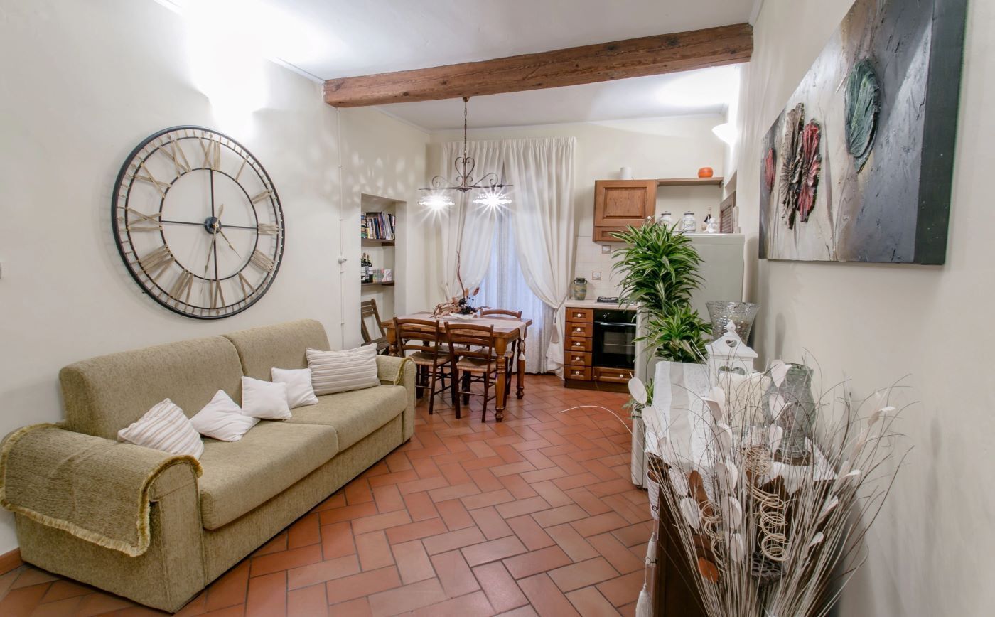 Un soggiorno con divano, tavolo da pranzo e sedie. Si nota un orologio a parete grande e un lampadario.