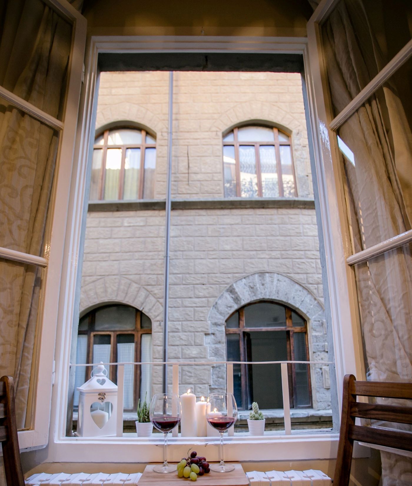 Una stanza con una grande finestra che si affaccia su un edificio in pietra, un tavolo con candele, bicchieri da vino e un cesto di frutta.