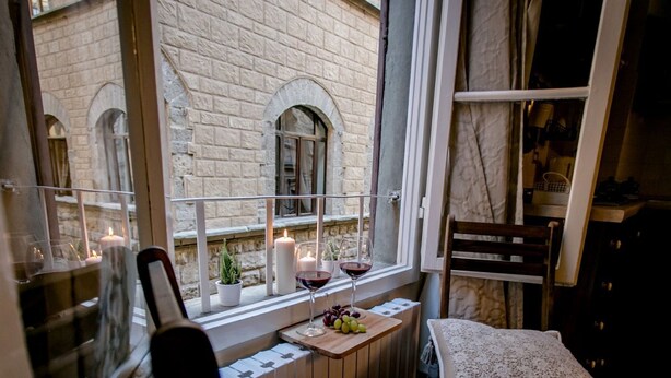 A cozy room with a wooden table set for two, a window with a view of a stone building, and a chair with a lace cover. - FLORENCE TOWER APARTMENT (Firenze)