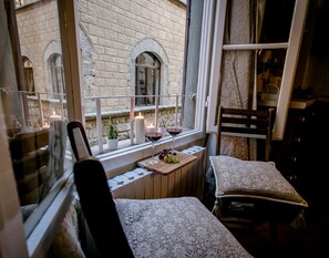 A cozy room with a wooden table set for two, a window with a view of a stone building, and a chair with a lace cover.