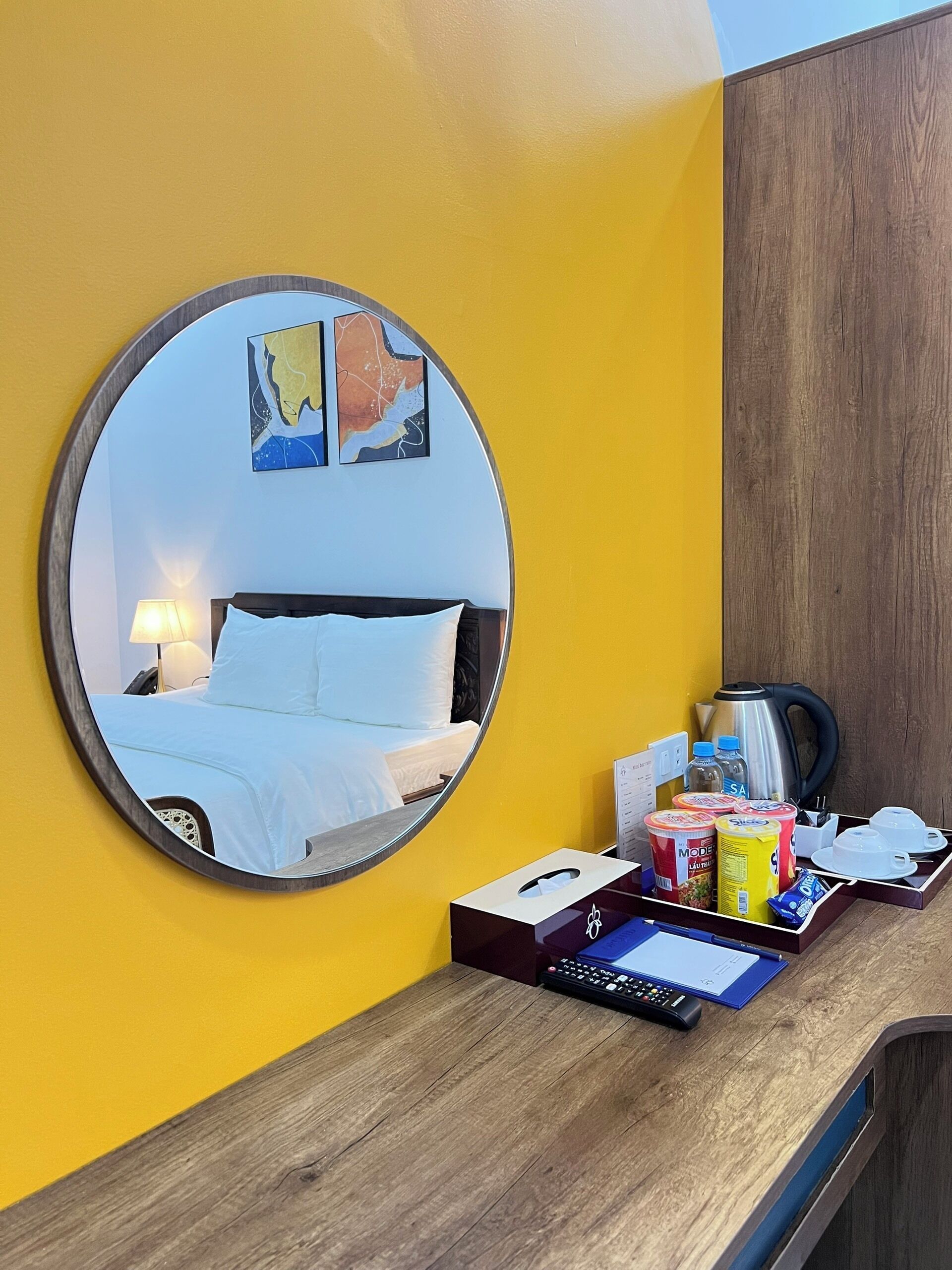 A hotel room with a round mirror reflecting a bed, a desk with a coffee maker, and various amenities.