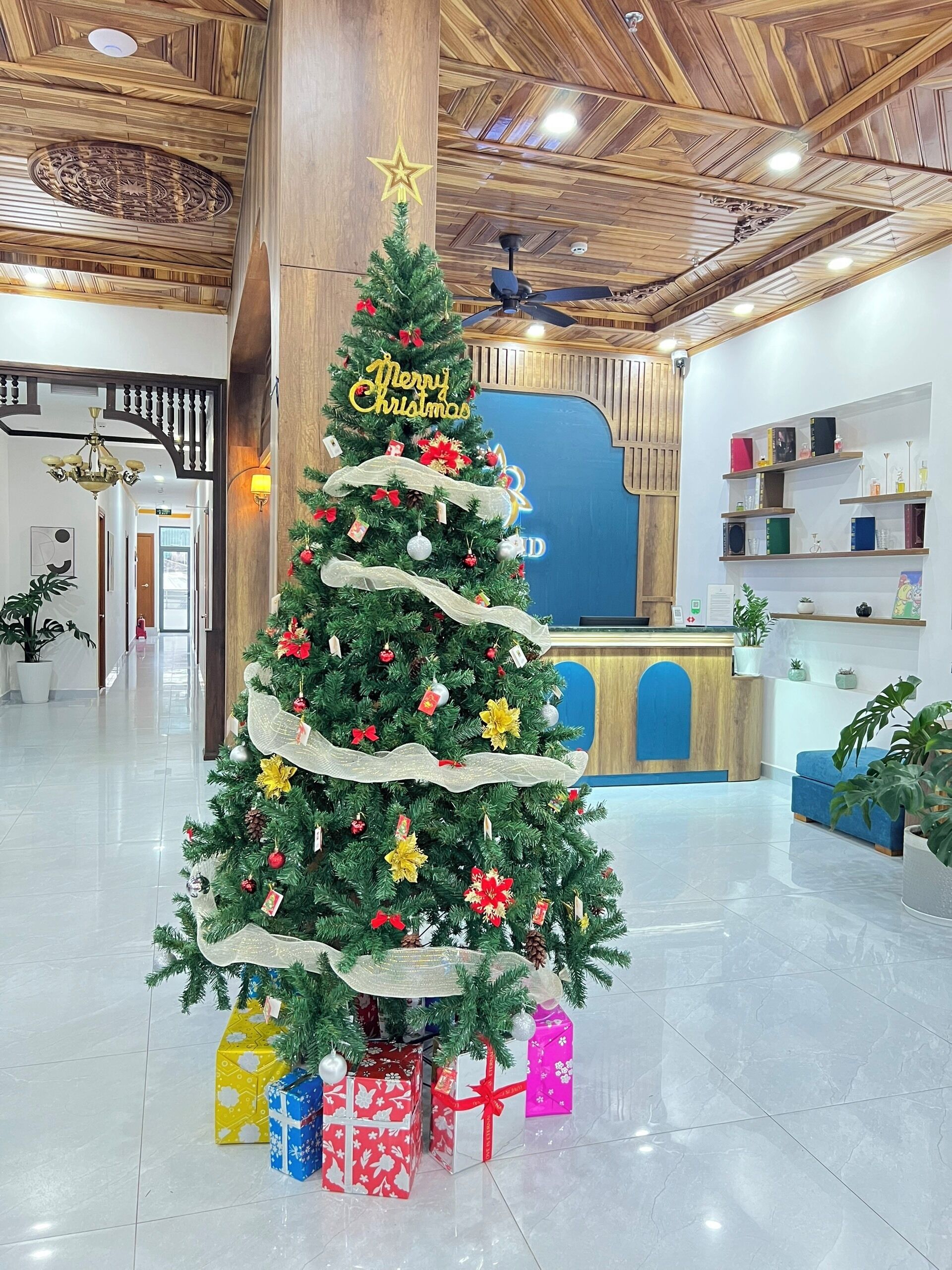 Un árbol de Navidad con adornos y un cartel que dice 'Merry Christmas', rodeado de cajas de regalo en un vestíbulo con techo de madera y un mostrador de recepción.