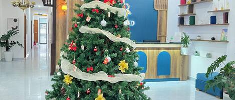 Un árbol de Navidad con adornos y un cartel que dice 'Merry Christmas', rodeado de cajas de regalo en un vestíbulo con techo de madera y un mostrador de recepción.
