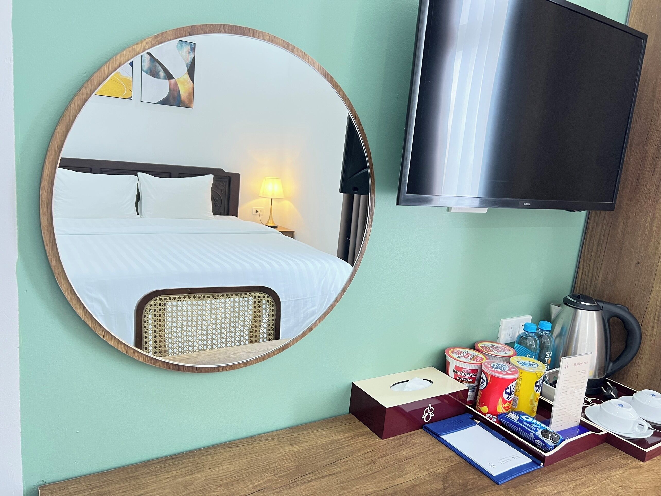 A hotel room with a bed, a round mirror, a television, and a table with amenities like a kettle, cups, and snacks.