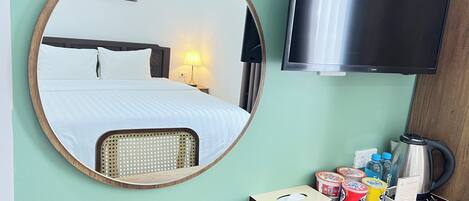 A hotel room with a bed, a round mirror, a television, and a table with amenities like a kettle, cups, and snacks.