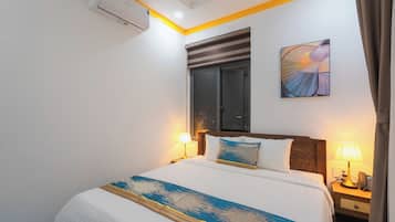 Habitación de hotel con cama, lámparas de noche, ventana con persianas, aire acondicionado y una obra de arte colgada en la pared.