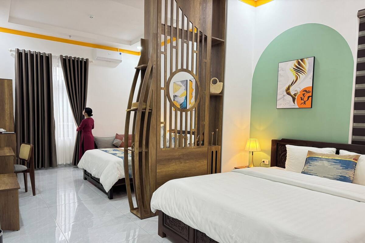 A hotel room with two beds, a wooden partition, a wall-mounted air conditioner, and a painting.