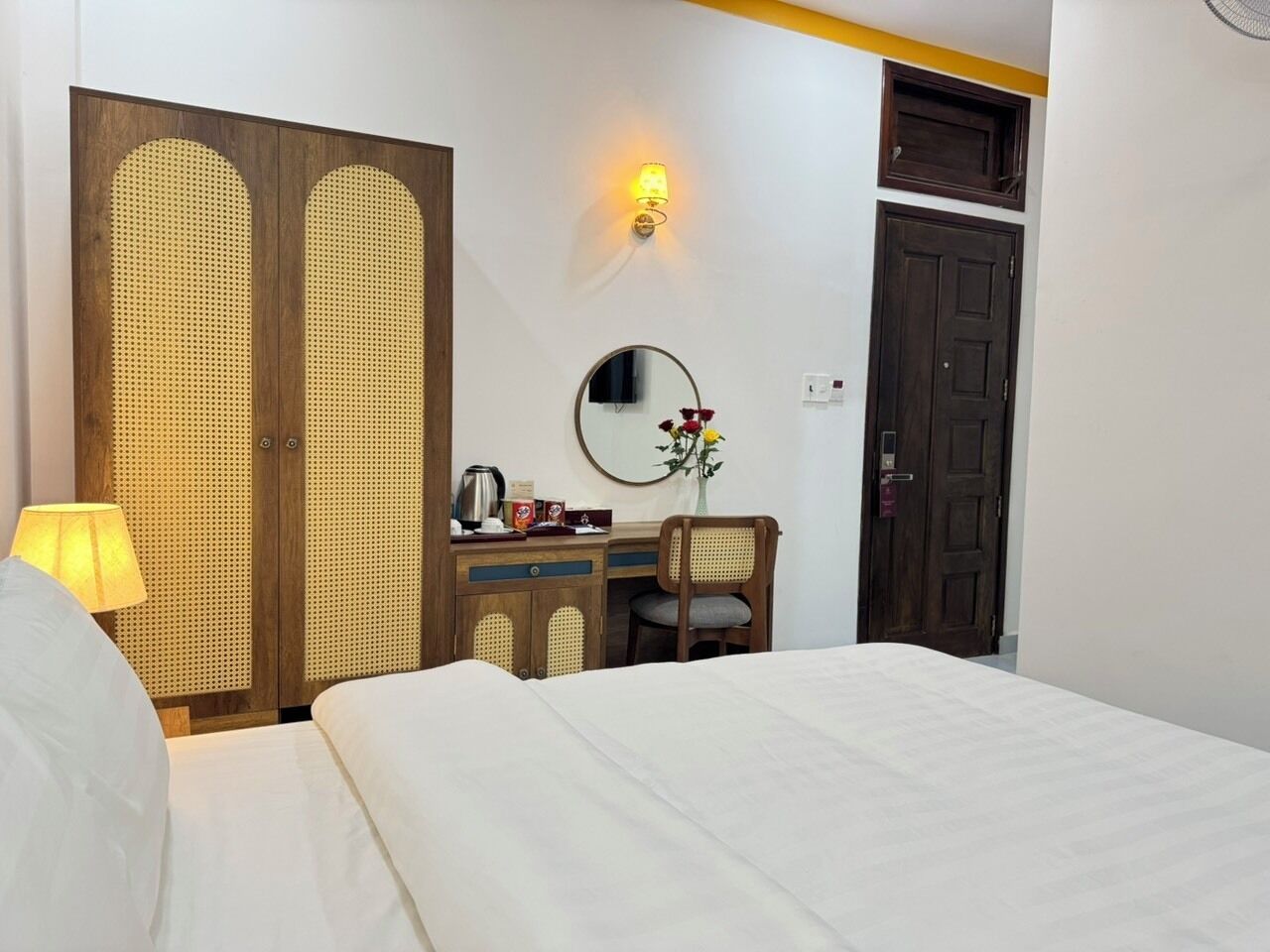 A hotel room with a bed, a desk with a chair, a mirror, a lamp, and a wardrobe.
